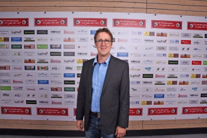 Mann im Anzug vor einer Sponsorwand mit vielen Logos, steht frontal für ein Presse-/Eventfoto und blickt in die Kamera.