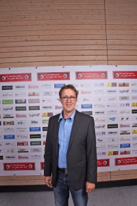 Mann im Sakko und Hemd steht vor einer Sponsorwand aus vielen Logos und Vereinsaufklebern; dient als Porträt im Eventkontext.