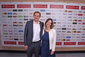 Zwei Personen posieren lächelnd vor einer Sponsorwand; formelle Kleidung, Hintergrundlogos dienen als Eventkulisse.