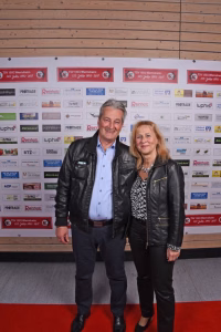 Zwei Personen auf rotem Teppich vor einer Sponsorwand, im Vordergrund gemeinsam posierend. Hintergrund dient als Veranstaltungs- und Werbefläche.