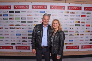 Zwei Personen stehen vor einer Sponsorwand und lächeln in die Kamera, aufgenommen bei einer Veranstaltung; Hintergrund mit vielen Logos und Vereinsplaketten.