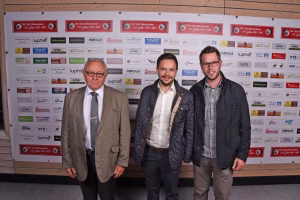 Drei Männer stehen vor einer Sponsorenwand; zwei im Anzug, einer in Jacke und Hemd, alle blicken in die Kamera.