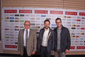 Drei Männer stehen nebeneinander und lächeln vor einer Sponsor-Wand mit vielen Logos, aufgenommen bei einer Veranstaltung.