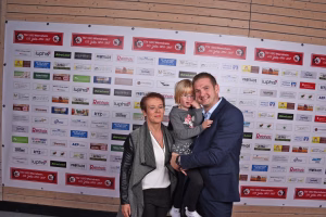 Familie vor einer Sponsorwand mit zahlreichen Logos und Jubiläumsbannern; Vater hält ein Kind im Arm und lächelt in die Kamera.