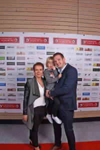 Familie steht auf rotem Teppich vor Sponsorenwand, Eltern halten ein Kleinkind; offizielle Fotomoment-Darstellung.