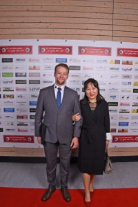 Zwei Personen im Anzug und Kleid posieren auf rotem Teppich vor einer Sponsorenwand, als offizielles Veranstaltungsfoto.