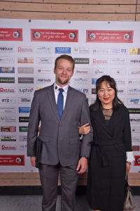 Zwei Personen im Anzug und Kleid stehen vor einer Sponsorwand mit Logos und halten für ein offizielles Event-Foto die Pose.