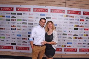 Zwei Personen posieren Schulter an Schulter vor einer Sponsorwand mit vielen Firmen-Logos; sie zeigen freundliche Gesichter für ein Foto.