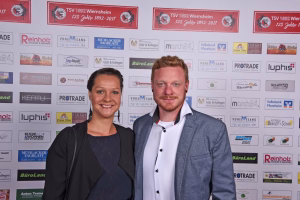 Zwei Personen vor einer Sponsorwand, lächeln in die Kamera; formelles Outfit für ein Presse- oder Veranstaltungsfoto.