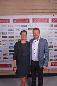 Zwei Personen posieren nebeneinander vor einer Sponsorwand mit bedruckten Logos und Jubiläumsbannern.