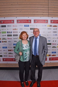 Zwei Personen posieren auf rotem Teppich vor einer Sponsorwand mit Logos, als offizielles Gruppenfoto bei Veranstaltung.