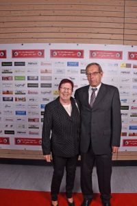 Zwei Personen posieren auf rotem Teppich vor einer Sponsorwand; das Foto dokumentiert den gemeinsamen Auftritt bei einer Veranstaltung.