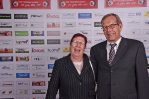 Zwei Personen posieren vor einer Sponsorwand mit Firmenlogos; Aufnahme für eine Veranstaltung, Blick nach vorne.