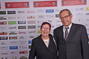 Zwei lächelnde Personen vor einer Sponsorwand mit vielen Logos und Schildern; Hintergrund dient der Veranstaltungspromotion.