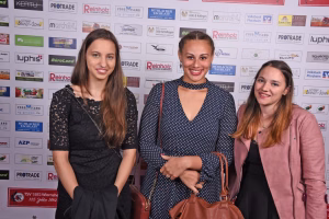 Drei lächelnde Frauen posieren vor einer mit Logos bedruckten Sponsorwand; scheinbar für ein Eventfoto oder eine Presseaufnahme.