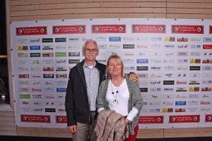 Zwei Personen posieren vor einer Sponsorwand; die Frau hält eine Jacke, beide stehen nah aneinander und lächeln bei einer Veranstaltung.