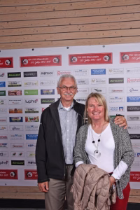 Zwei Personen posieren lächelnd vor einer Sponsorwand mit vielen Logos und Texten; die Szene zeigt ein offizielles Gruppenfoto bei einer Veranstaltung.