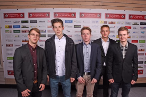 Fünf Personen in Anzügen und Hemden posieren vor einer Sponsorwand mit Logos, für ein Gruppenfoto bei einem Event.
