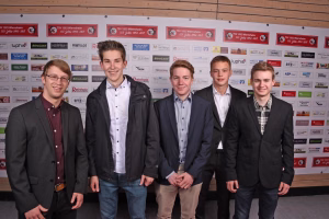 Fünf Personen in Anzügen und Hemden vor einer bedruckten Sponsorenwand, Fotocall bei einer Veranstaltung.