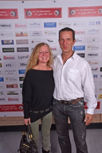 Zwei Personen stehen vor einer Sponsorwand; sie posieren lächelnd für ein Erinnerungsfoto bei einer Veranstaltung.