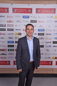 Mann im Anzug posiert vor einer bedruckten Sponsorwand bei einer Veranstaltung und präsentiert sich für ein Pressefoto.