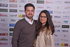 Zwei Personen lächeln vor einer Sponsorwand mit Logos; gemeinsames Portrait für eine Veranstaltung oder Pressepräsenz.