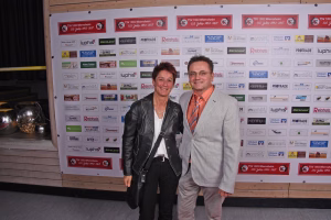 Zwei Personen vor einer Sponsorwand mit Logos und Jubiläumsaufdrucken; posieren für ein Gruppenfoto bei einer Veranstaltung.