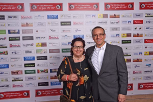 Zwei Personen stehen vor einer Sponsorwand und lächeln in die Kamera; im Vordergrund gemeinsames Foto bei Veranstaltung.