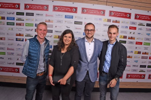 Vier Personen stehen gemeinsam vor einer Sponsorenwand und lächeln in die Kamera, vermutlich für eine Veranstaltung oder Preisübergabe.