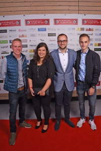 Vier Personen stehen nebeneinander vor einer Sponsorwand und posieren in Business- und Freizeitkleidung auf rotem Teppich.