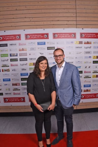Zwei Personen posieren auf rotem Teppich vor einer Sponsorwand mit vielen Logos und Jubiläums-Aufdrucken.