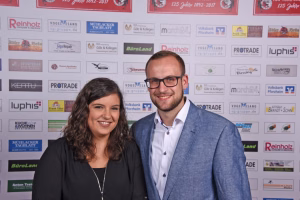 Zwei Personen lächeln nebeneinander vor einer Wand mit Firmen-Logos; wirkt wie ein gemeinsames Veranstaltungs- oder Pressefoto.