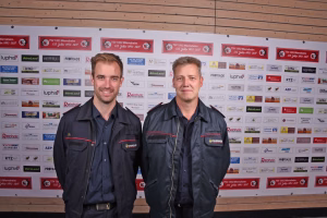 Zwei Feuerwehrmänner stehen vor einer Sponsorwand und lächeln in die Kamera, getragenen Dienstjacken mit Abzeichen und Schutzfunktion.