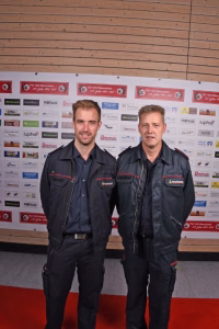 Zwei Feuerwehrleute in dunkler Einsatzkleidung posieren vor einer Sponsorwand mit Logos; Hintergrund dient als Veranstaltungs- und Kontextanzeige.
