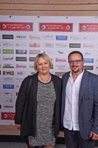 Zwei Personen posieren vor einer Wand mit Sponsorlogos, lächeln in die Kamera bei einer Veranstaltung.