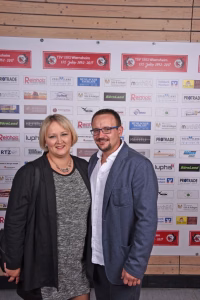 Zwei Personen stehen nebeneinander vor einer mit Vereins- und Sponsorlogos bedruckten Wand, lächeln in die Kamera.