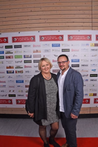 Zwei Personen posieren vor einer Sponsorwand mit zahlreichen Logos, im Vordergrund, für ein Eventfoto.