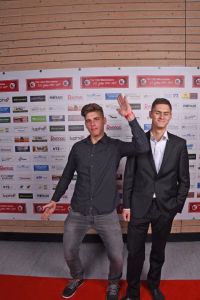 Zwei Männer posieren auf rotem Teppich vor einer Sponsorwand und winken in die Kamera. Holzwand im Hintergrund, sportliche Veranstaltung.