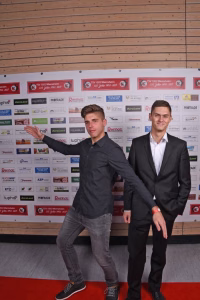 Zwei Männer posieren auf einem roten Teppich vor einer Sponsorwand aus bedruckten Bannern, eine Person zeigt lässig mit der Hand.
