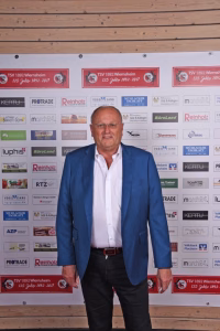 Mann im Anzug steht vor Sponsorenwand mit Logos, unter Holzrahmen; Blick in die Kamera, Fotoposition bei Veranstaltung.