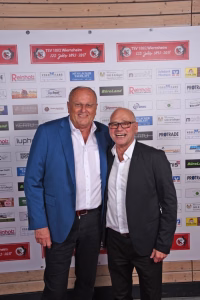 Zwei Männer stehen lächelnd nebeneinander vor einer Sponsorwand mit Logos und Texten bei einem Event, zur Begrüßung und Darstellung.