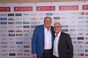 Zwei Männer stehen vor einer Wand mit vielen Logos und Schildern, im Anzug, lächelnd; wirkt wie Teilnehmerfoto bei einer Veranstaltung.