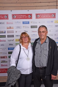 Zwei Personen vor einer Wand mit Sponsor-Aufklebern, eine Frau hält einen Arm um die Schulter des Mannes; gemeinsames Gruppenfoto.