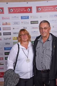 Zwei Personen stehen vor einer Sponsorenwand mit Firmenlogos; sie posieren für ein Eventfoto im Indoor-Eingangsbereich.