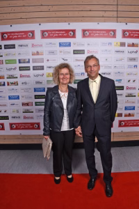 Zwei Personen stehen nebeneinander auf rotem Teppich vor einer Sponsorwand, lächeln in die Kamera; formelles Event-Foto.