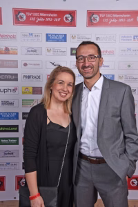 Zwei Personen stehen lächelnd vor einer Sponsorwand mit Logos und Jubiläumsbändern, um einen offiziellen Empfang/Foto zu dokumentieren.