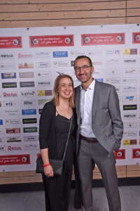 Zwei Personen posieren lächelnd an einer Sponsorwand mit vielen Logos und Jubiläums-Aufklebern, im Hintergrund eine Holzverkleidung.