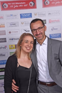Zwei Personen lächeln in Anzug- und Businesskleidung vor einer Wand mit Sponsorenlogos bei einem Event.