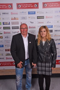 Zwei Personen stehen nebeneinander vor einer Sponsorwand, lächeln in die Kamera; festliches Rahmenfoto bei einem Event.