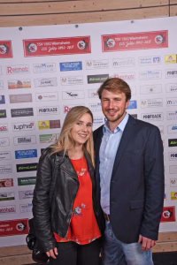 Zwei Personen lächeln gemeinsam vor einer Sponsorwand; die Logos bilden den Hintergrund, sie dienen als Anlass- und Fotokulisse.